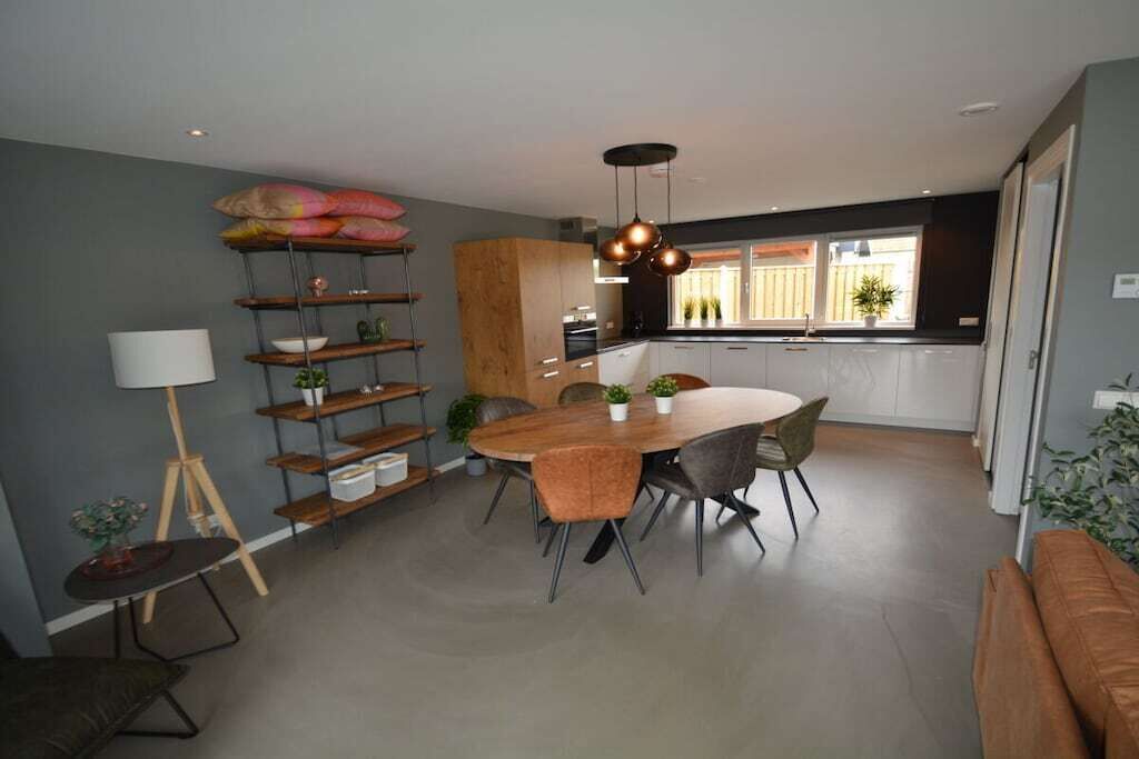 A modern kitchen with a dining area, featuring a round table with chairs, a pendant light, and a shelving unit with plants.
