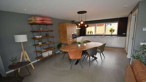 A modern kitchen with a dining area, featuring a round table with chairs, a pendant light, and a shelving unit with plants. - Holiday apartment with 1 bedroom (Serooskerke)