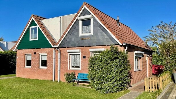 Une maison à deux étages avec un toit vert et gris, une façade en briques et un petit jardin devant, agrémenté d’un banc et d’un arbuste.