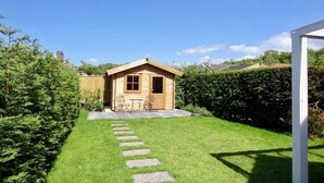En træhytte med en lille terrasse, omgivet af en velplejet græsplæne og en hæk.