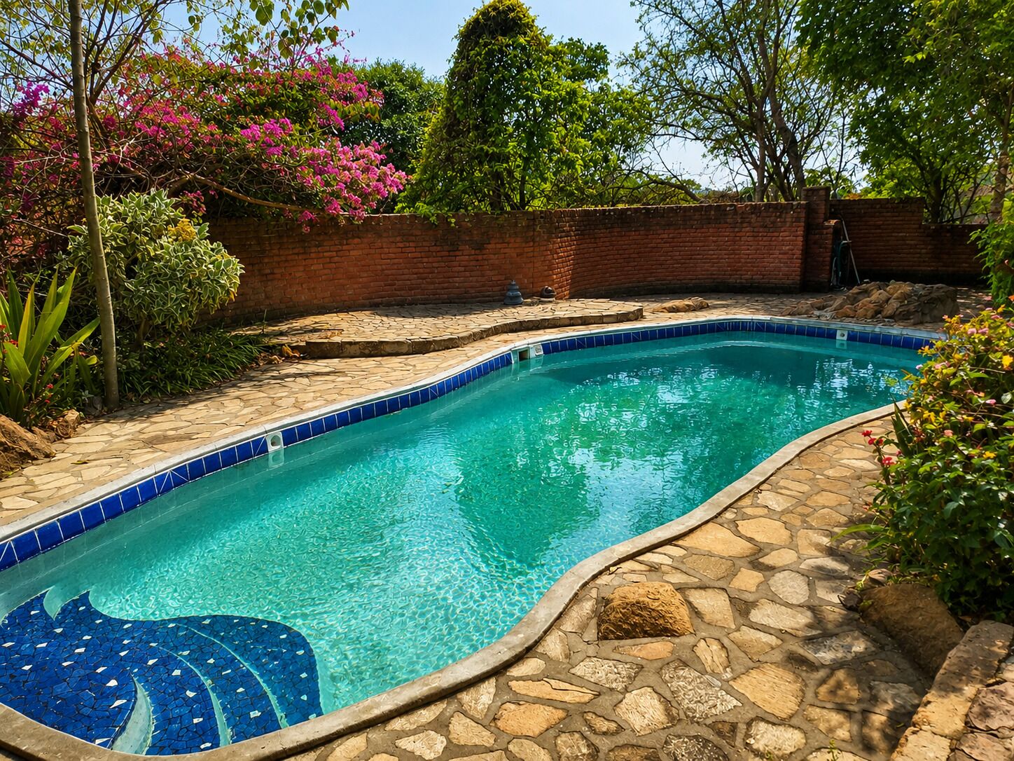 Una piscina con borde de azulejos y un foso de piedra, rodeada de follaje y un muro de ladrillo.
