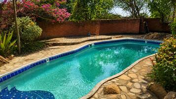 Una piscina con borde de azulejos y un foso de piedra, rodeada de follaje y un muro de ladrillo.