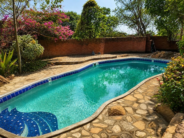 A swimming pool with a tiled edge and a stone border, surrounded by greenery and a brick wall. - Pagomo Lodges (Harare)