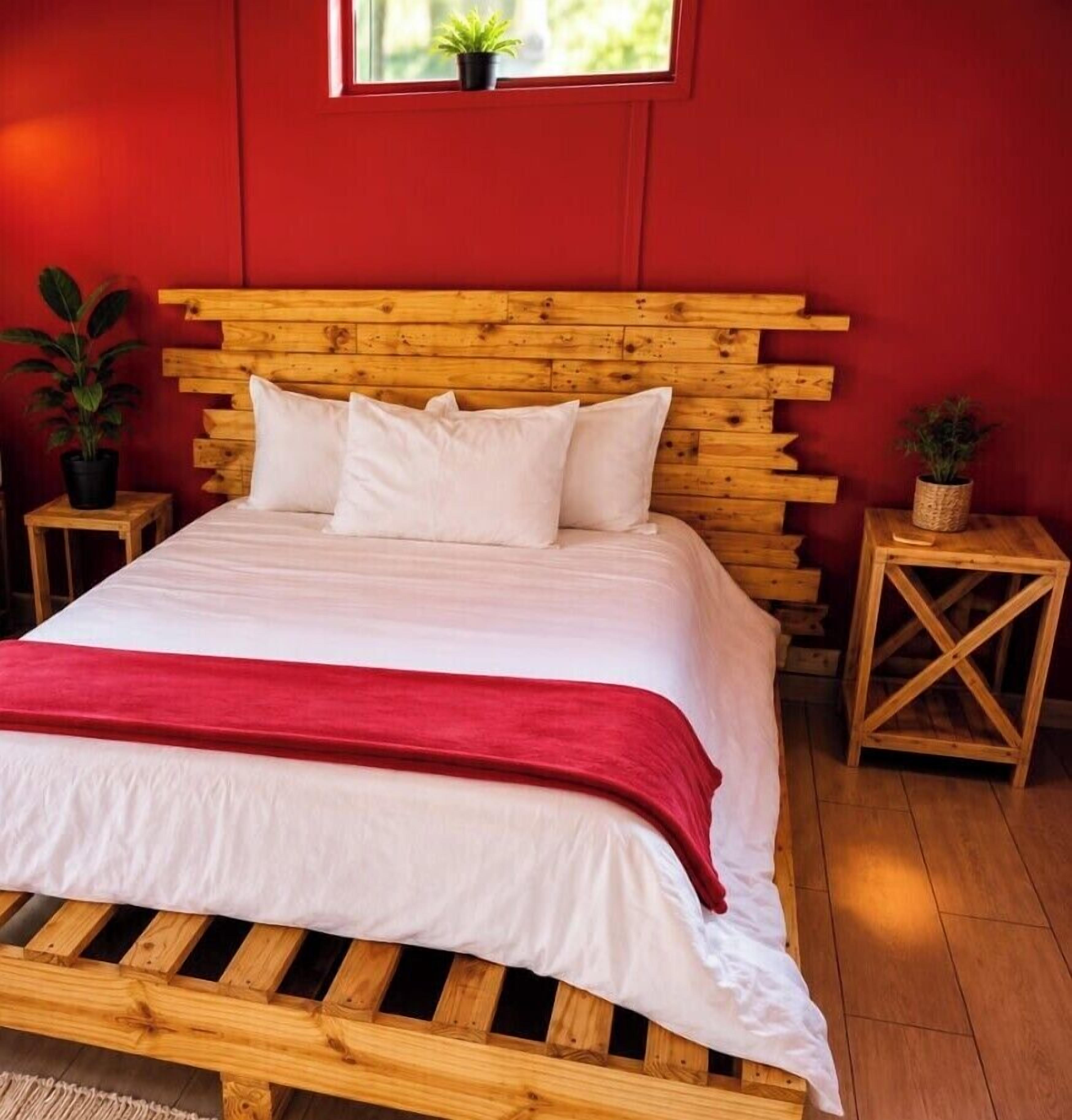 Ein Bett mit hölzernem Kopfteil, weißer Bettwäsche und einer roten Tagesdecke. Es gibt zwei Nachttische aus Holz, jeweils mit einem Blumentopf.