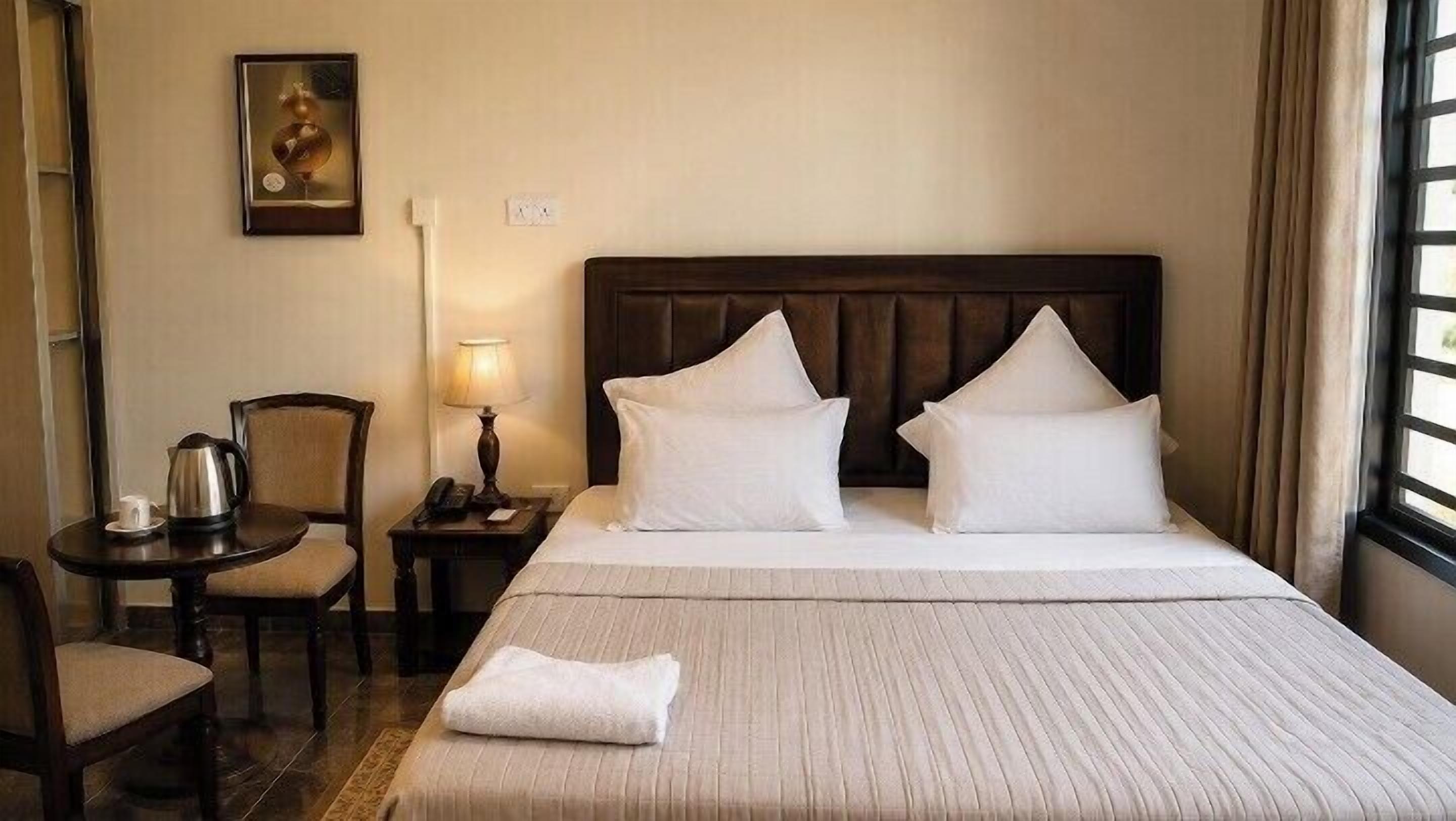 Habitación de hotel con una cama grande, dos almohadas, una mesita de noche con una lámpara, una silla y un cuadro en la pared.