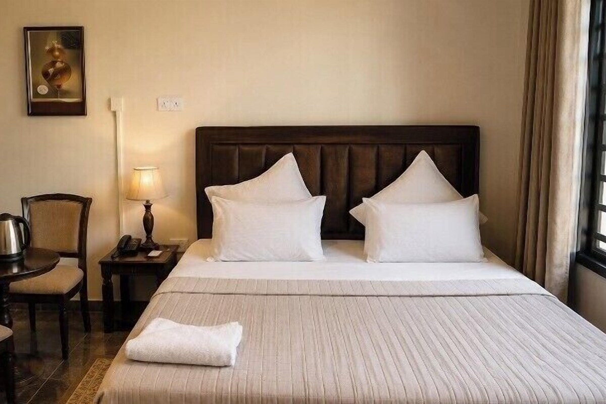 A hotel room with a large bed, two pillows, a bedside table with a lamp, a chair, and a painting on the wall.