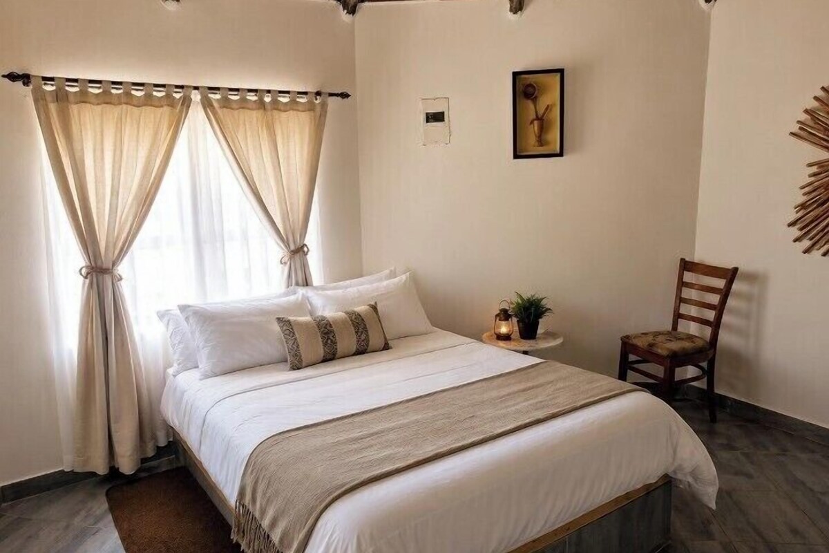 A bedroom with a bed, curtains, a chair, a small table with a plant, and a wall-mounted picture.