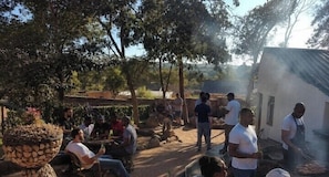 An outdoor gathering with people sitting and standing, a cooking area with smoke, and a rustic building in the background.
