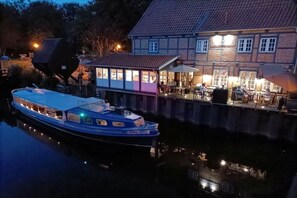 Ein Boot liegt an einem Flussufer, mit einem Gebäude mit großen Fenstern und Außenplätzen.