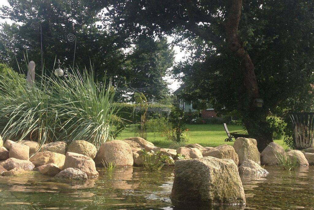 Un étang bordé de rochers et d’herbe, entouré d’arbres, avec une maison visible au loin.