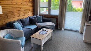 A cozy room with a wooden wall, a grey sofa, a white armchair, and a small wooden table with a remote control.