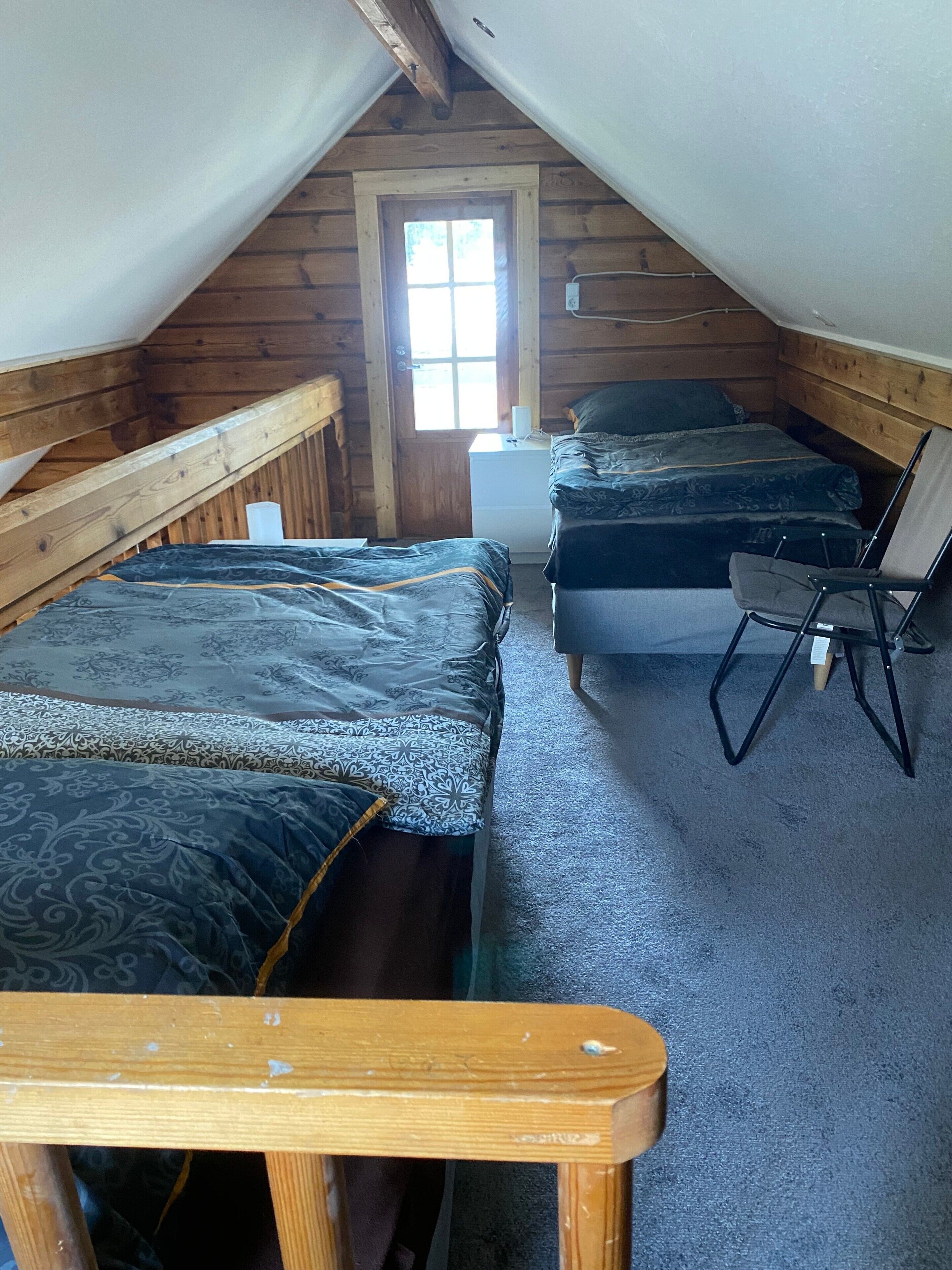 A small, cozy attic bedroom with two beds, a window, and a wooden chair.