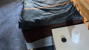 Quarto pequeno com cama de solteiro, teto inclinado, janela e mesa com controle remoto.