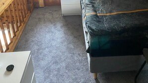 A small, cozy room with a wooden ceiling, a single bed with a patterned bedspread, a nightstand with a lamp, and a door leading outside.