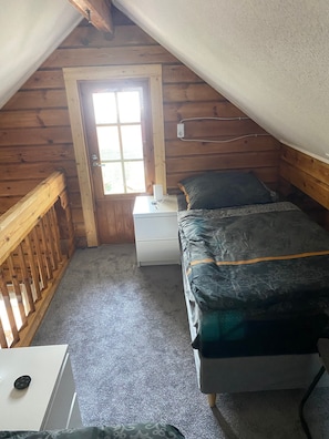 A small, cozy room with a wooden ceiling, a single bed with a patterned bedspread, a nightstand with a lamp, and a door leading outside.