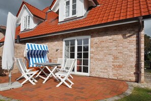 A brick house with a red-tiled roof, a large window, and a patio with a table and chairs. - Holiday home with 2 bedrooms (St.Peter-Ording)