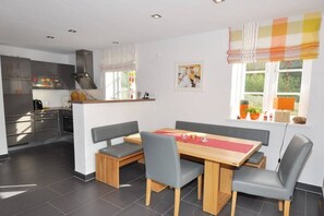 A modern kitchen with built-in appliances, a dining area with a wooden table and grey upholstered chairs, and a window with a view of greenery. - Holiday home with 2 bedrooms (St.Peter-Ording)