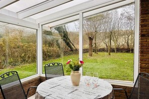 Une véranda avec une table et des chaises, un vase rempli de fleurs et une vue sur un espace verdoyant avec des arbres.