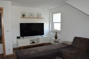 Een woonkamer met een flatscreen televisie op een witte standaard, een grijze bank en een bijzettafeltje met een vaas.
