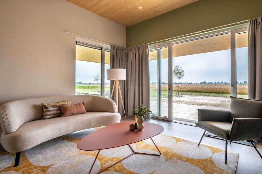 Un salon moderne avec un canapé, un fauteuil, une table basse et une lampe. Une grande fenêtre, ornée de rideaux, offre une vue sur l’extérieur.