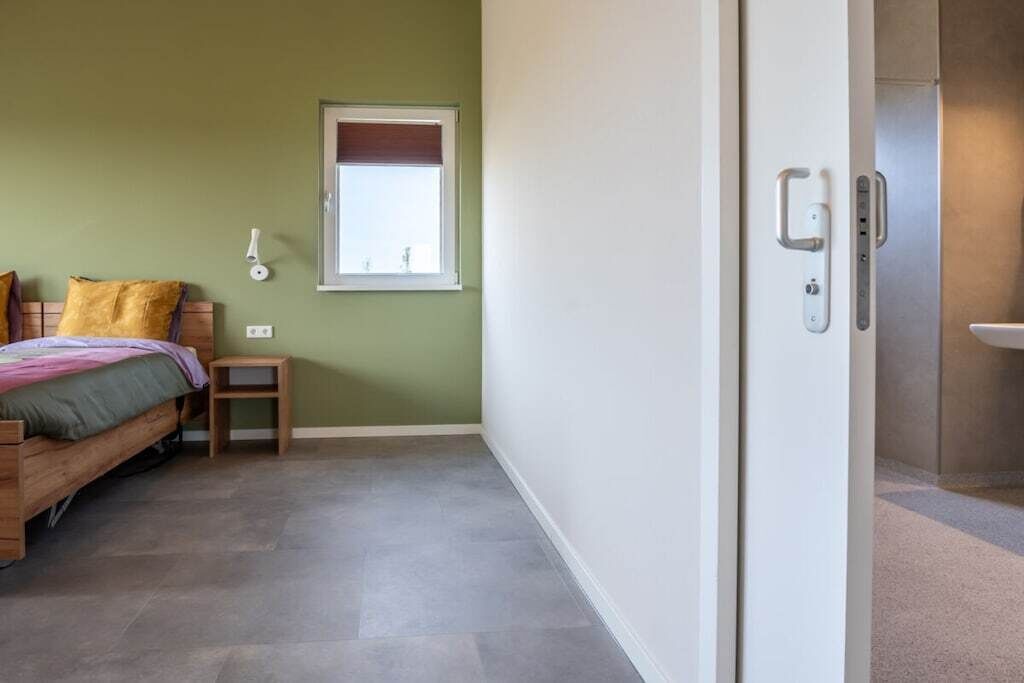 A bedroom with a bed, a small bedside table, a wall-mounted light, and a window with a view.