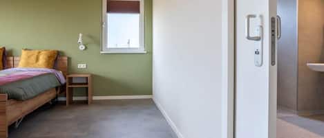 A bedroom with a bed, a small bedside table, a wall-mounted light, and a window with a view.