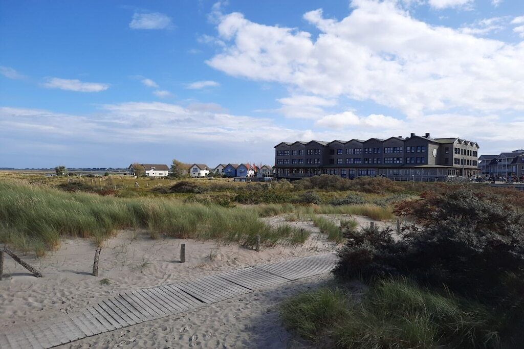 Ein Küstenhotel mit Sandstrand und Dünen im Vordergrund, sowie ein modernes Gebäude mit vielen Fenstern im Hintergrund.
