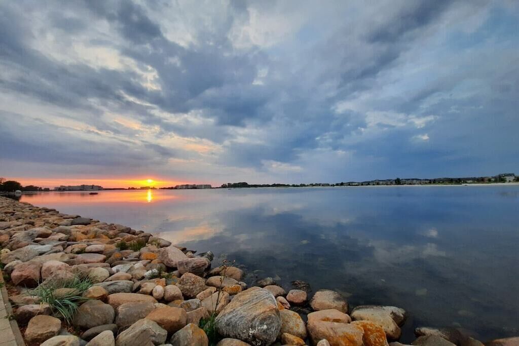Ein ruhiger See bei Sonnenuntergang mit einer felsigen Uferlinie und Gebäuden in der Ferne.