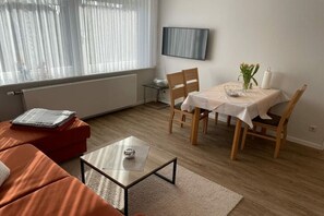 A modern living room with a sofa, a coffee table, a dining table with chairs, a TV mounted on the wall, and a window with sheer curtains.