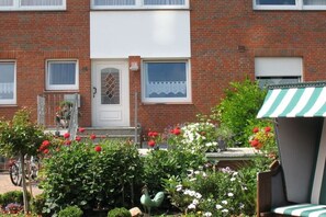 Una casa de ladrillo con una puerta blanca, un pequeño porche con barandilla y un jardín con diversas plantas y flores.