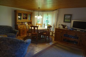Ein Wohnzimmer mit Esstisch, Stühlen, einer Couch, Holzschränken und einem Fernseher.