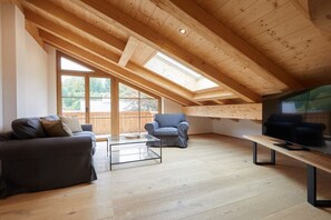 Ein modernes Loft mit schrägem Dach, großen Fenstern, einer Couch, einem Sessel und einem Fernseher auf einem Holztisch.