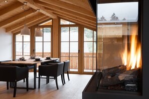 Ein Essbereich mit Holztisch und -stühlen, großen Fenstern und einem Kamin, der durch ein Glasfenster sichtbar ist.