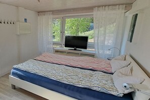 Ein Schlafzimmer mit einem Bett, einem Fernseher auf einem Schrank, einem Fenster mit Vorhängen und einem an der Wand befestigten Lichtschalter.