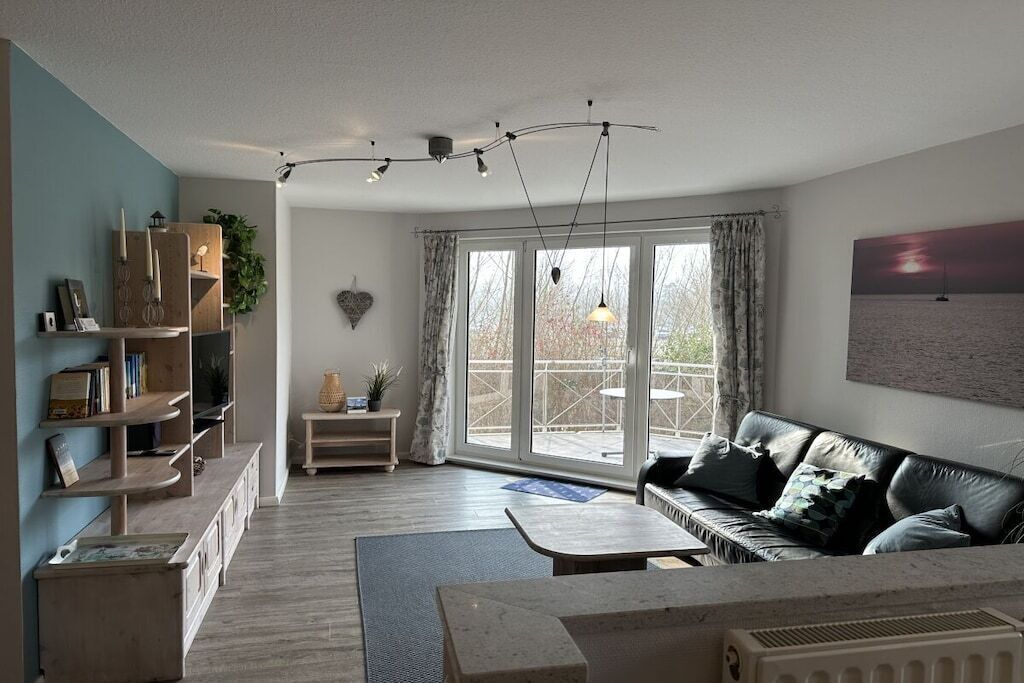 Een moderne woonkamer met een bank, salontafel en een open haard. Er is een groot raam met gordijnen, een boekenkast en een plant.