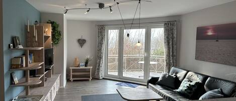Een moderne woonkamer met een bank, salontafel en een open haard. Er is een groot raam met gordijnen, een boekenkast en een plant.