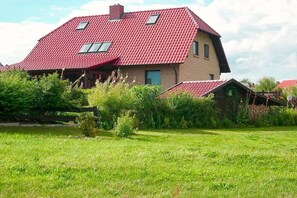 A two-story house with a red roof, surrounded by a grassy field and dense shrubbery. - Holiday apartment with 1 bedroom (Malchow)