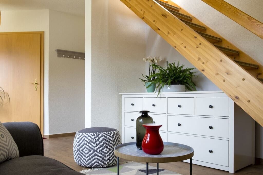A room with a white dresser, a round table with a red vase, a patterned ottoman, and a wooden staircase.