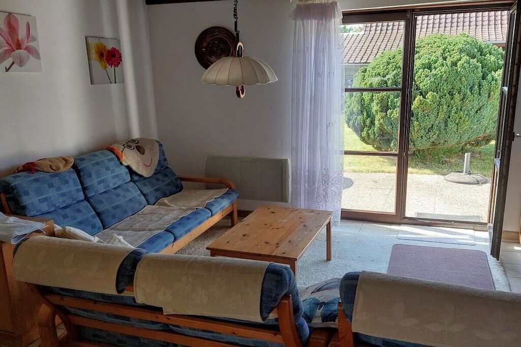 Una sala de estar con un sofá, una mesa de centro de madera y vista a un arbusto verde a través de un ventanal.