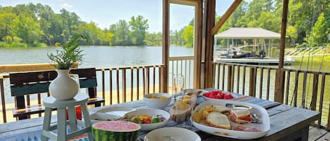 A wooden deck with a table set for a meal, overlooking a lake with a gazebo in the background.