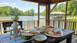 Un terrazzo in legno con un tavolo apparecchiato per un pasto, con vista su un lago e un gazebo sullo sfondo.