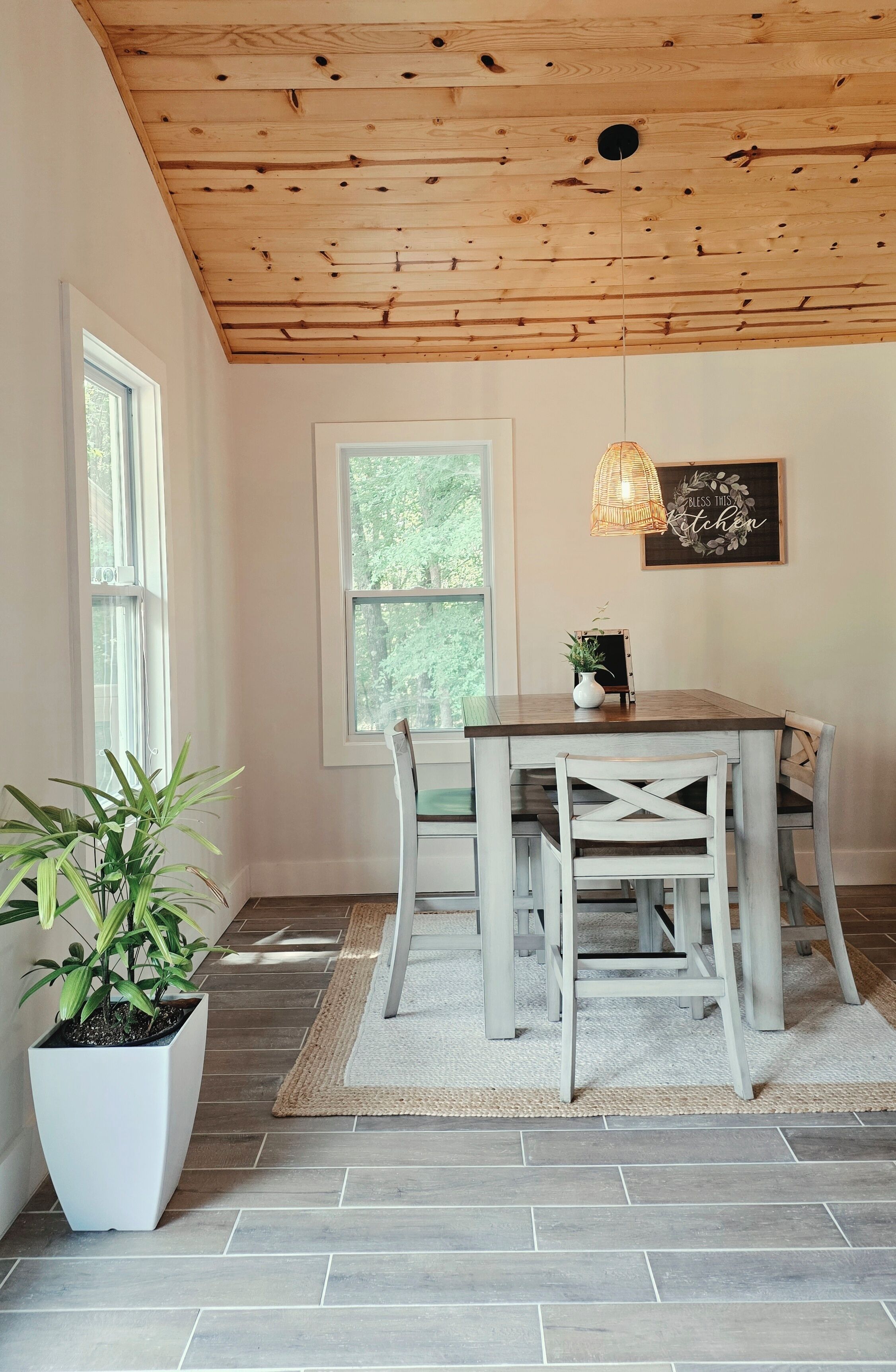 A dining area with a wooden ceiling, a table with chairs, a potted plant, and a window with a view of trees.