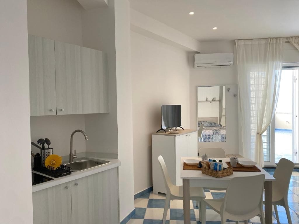 Cocina compacta con pileta, pequeño refrigerador y comedor con mesa y sillas.