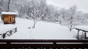 A wooden cabin with a snow-covered roof, surrounded by snow-laden trees and a wooden deck.