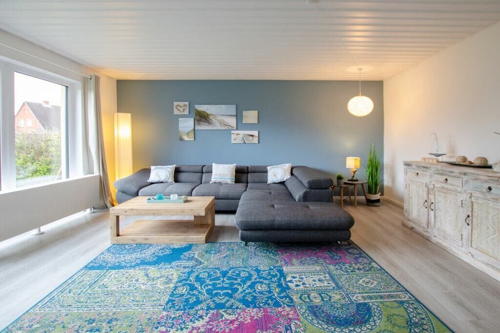 Een moderne woonkamer met een grijze modulaire bank, een houten salontafel en een kleurrijk vloerkleed. Er staat een kast met decoratieve objecten en een lamp op een bijzettafeltje.
