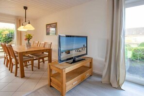 Een eetruimte met een houten tafel en stoelen, een televisie op een houten dressoir, en een raam met gordijnen.