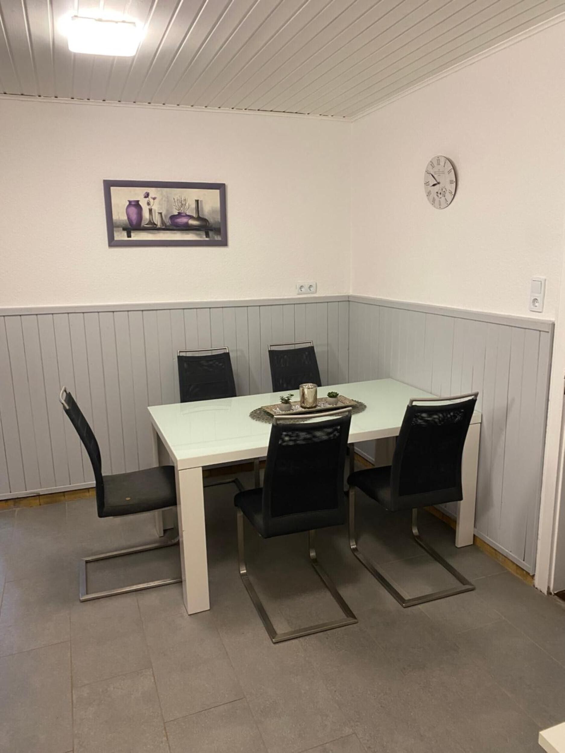 Un comedor con una mesa blanca, sillas negras, un reloj en la pared y un cuadro colgado sobre la mesa.