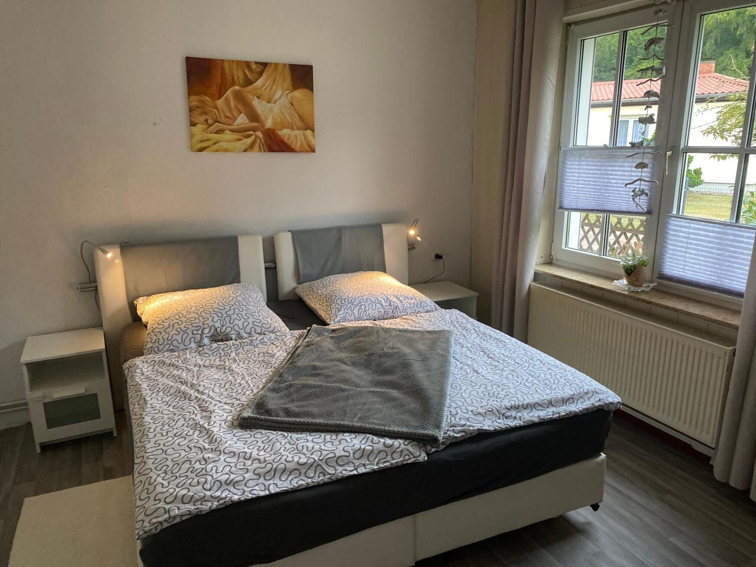 Un dormitorio con cama, mesitas de noche, una ventana con vistas y un cuadro en la pared.