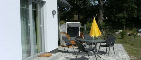 Un patio exterior con mesa y sillas, un paraguas amarillo y una silla de mimbre con cojín negro.