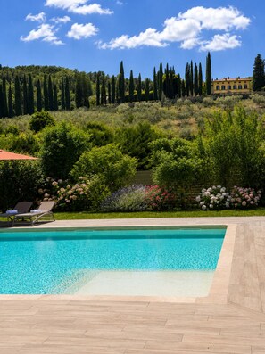 A swimming pool with lounge chairs, surrounded by lush greenery and a hill with cypress trees. - Vrbo Property (Arezzo)